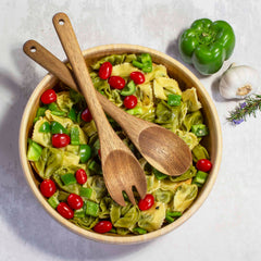 An acacia wood bowl from Totally Bamboo® is filled with tortellini pasta, diced green bell pepper, and cherry tomatoes, and is accompanied by a TB Home® Acacia Wood Salad Serving Set, 12". Next to the bowl are a whole green bell pepper, a garlic bulb, and a rosemary sprig on a light background.