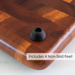 Close-up of one corner of the Rock & Branch® Acacia Charcuterie Board with Cheese Slicer by Totally Bamboo, highlighting a black non-skid foot beneath it. The elegant checkered surface, crafted from light and dark wood, is prominently featured. Text on the image reads, "Includes 4 Non-Skid Feet.