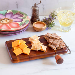 A Totally Bamboo Rock & Branch® Acacia Charcuterie Board with included cheese slicer, featuring sliced cheddar and pepper jack cheese, rye crackers, colorful plates, a pepper mill, rosemary sprigs, and a glass of iced lemonade, adorns the marble countertop.