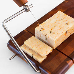 The Rock & Branch® Acacia Charcuterie Board with Cheese Slicer by Totally Bamboo effortlessly slices through a block of herb-infused cheese. Two perfectly cut slices rest elegantly beside the block.