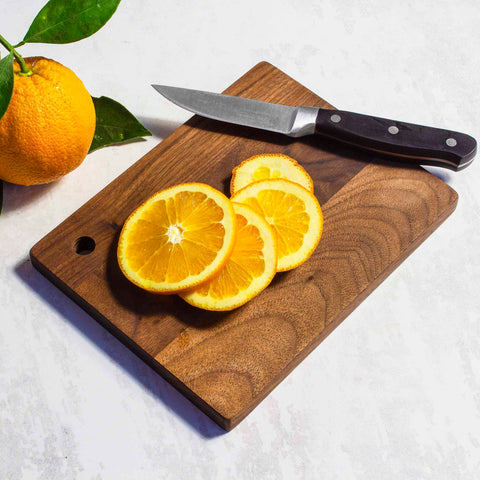 Sliced oranges rest on the TB Home® Walnut Wood Small Cutting Board, 8" x 6-3/8", by Totally Bamboo, with a knife. An unsliced orange, adorned with green leaves, sits beside the board on a white surface, adding rustic elegance to the scene.