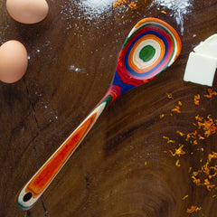 A vibrant Totally Bamboo Baltique® Marrakesh Collection Cooking Spoon, 12-1/2", crafted from colored birch with a circular pattern, rests on a dark wooden surface. Surrounding it are scattered eggs, a cube of butter, grated orange zest, and a dusting of flour.