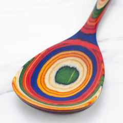 The Totally Bamboo Baltique® Marrakesh Collection Cooking Spoon, 12-1/2", crafted from colored birch, showcases swirling rainbow patterns on a white marble surface. Its vibrant design features concentric circles in blue, red, yellow, green, and other colors for an eye-catching appearance.