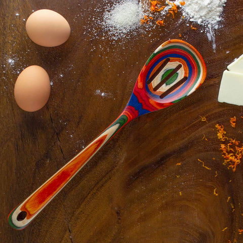 A lively, striped slotted spoon from the Baltique® Marrakesh Collection by Totally Bamboo rests on a wooden surface, accompanied by two eggs, scattered flour, sugar, grated orange zest, and a block of butter. This colorful kitchen tool is both visually striking and safe for use with non-stick cookware.