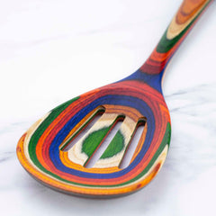 This vibrant and colorful slotted spoon, part of the Totally Bamboo Baltique® Marrakesh Collection, showcases swirling patterns of red, orange, green, blue, and beige against a white marble background. The design beautifully covers the entire 12-1/2" utensil, ensuring it's non-stick safe while creating a striking visual effect.