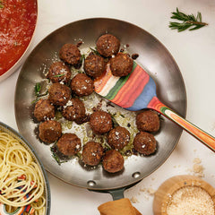 A skillet filled with cooked meatballs garnished with herbs and cheese rests next to a bowl of tomato sauce and a plate of spaghetti. The vibrant touch is provided by the non-stick safe, colorful Baltique® Marrakesh Collection Spatula by Totally Bamboo, alongside a small bowl of grated cheese.