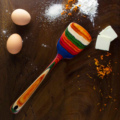 A vibrant spatula from the Totally Bamboo Baltique® Marrakesh Collection, measuring 12-1/2 inches, rests on a dark wooden surface, surrounded by two eggs, a small pile of flour, three pieces of butter, and some orange zest.