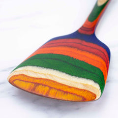 The vibrant Baltique® Marrakesh Collection Spatula by Totally Bamboo, measuring 12-1/2 inches, elegantly sits on the marble surface, displaying colorful stripes of blue, green, red, orange, and cream. Expertly designed to be non-stick safe, it brings a lively touch to any kitchen setting.