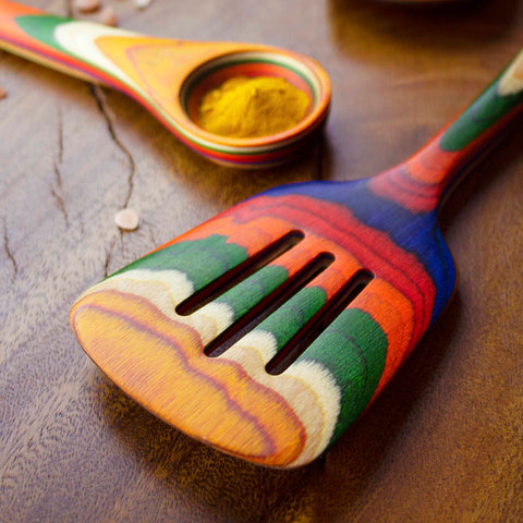 Part of the Totally Bamboo's Baltique® Marrakesh Collection, this vibrant kitchen utensil set includes a colorful wooden slotted spatula with rainbow-like patterns. The accompanying spoon delicately holds a hint of yellow spice, providing contrast to these lively tools resting on a wooden surface.