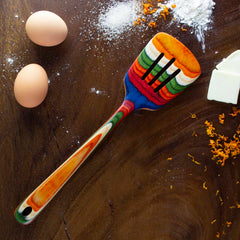 A colorful Baltique® Marrakesh Collection Slotted Spatula, 12-1/2" by Totally Bamboo is placed on a wooden surface, accompanied by two eggs, a pat of butter, orange zest, and scattered flour.