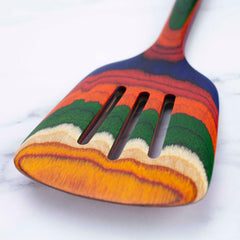 As part of the Baltique® Marrakesh Collection by Totally Bamboo, this colorful kitchen utensil showcases a slotted spatula design with vibrant horizontal stripes in orange, red, green, blue, and natural wood tones, elegantly displayed on a white marble surface.