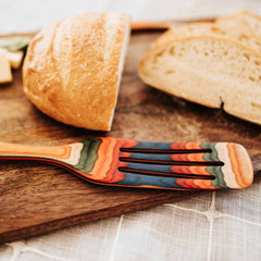 A vibrant spurtle from the Baltique® Marrakesh Collection by Totally Bamboo, featuring a rainbow-like pattern, rests on a wooden board beside sliced rustic bread. Its colorful birch design beautifully complements the light, textured fabric in the background.