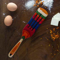 A brightly colored Baltique® Marrakesh Collection Spurtle, 13" from Totally Bamboo rests on a dark wooden surface, accompanied by two eggs, a dash of flour, a few cubes of butter, and some orange zest.