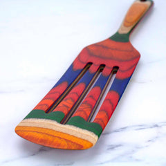 A vibrant wooden spatula from the Totally Bamboo Baltique® Marrakesh Collection Spurtle, 13", with layers of red, blue, green, and yellow hues, rests on a white marble surface. This versatile tool is made from colored birch for both style and functionality.