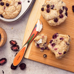 A wooden board showcases a halved blueberry scone adorned with butter. Nearby, whole scones rest alongside a Totally Bamboo Baltique® Marrakesh Collection Spreader Knife, 8", adding flair to the scene. Fresh berries complement the arrangement, and a muffin tin overflows with more delectable scones.