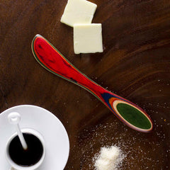 An 8-inch Baltique® Marrakesh Collection Spreader Knife by Totally Bamboo, known for its vibrant wooden design, rests on a dark table beside two small squares of butter. Below, a white cup of black coffee accompanied by a white stirrer and a dash of sugar evokes images of colorful marketplaces.