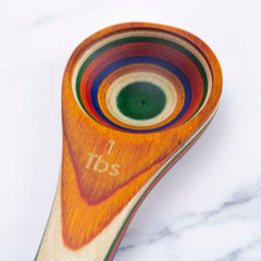 A product from the Marrakesh Collection, the Baltique® 2-in-1 Measuring Spoon by Totally Bamboo, showcases a vibrant multicolored design of concentric circles on colored birch. The spoon has a smooth finish with "1 TBS" engraved on its handle and is elegantly displayed against a white, marble-like background.