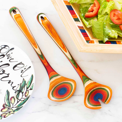 A pair of the vibrant Baltique® Marrakesh Collection Salad Serving Set, 11-3/4" by Totally Bamboo rests on a marble surface next to a salad bowl filled with lettuce and tomato slices. Nearby, a decorative plate adorned with the words "Olive Oil" and painted olive branches captures the essence of Morocco's colorful marketplaces.