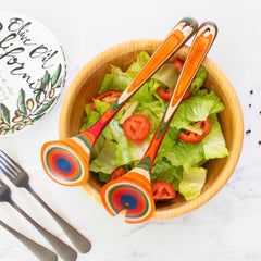 A wooden bowl filled with a green salad and tomato slices is topped with the vibrant Totally Bamboo Baltique® Marrakesh Collection Salad Serving Set, 11-3/4". Nearby, a decorative plate featuring floral designs and black forks rests on a marble surface, evoking the colorful marketplaces of Morocco.