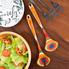 A wooden table displays a green salad with tomato slices in a light-colored bowl, enhanced by the vibrant design of the Baltique® Marrakesh Collection Salad Serving Set, 11-3/4" from Totally Bamboo, evoking the colorful essence of Moroccan marketplaces. Additionally, three dark forks are present alongside a white plate adorned with decorative text that is partially visible at the top.