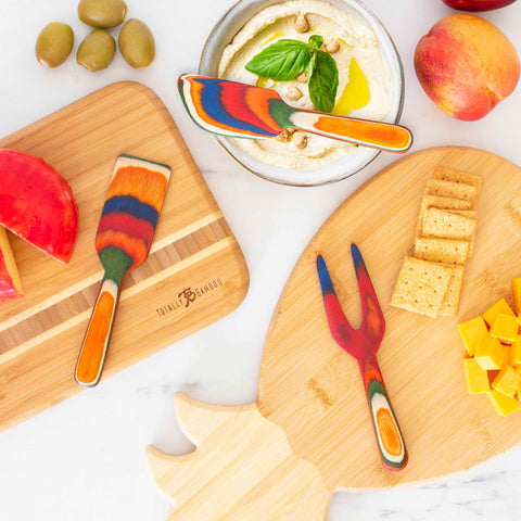 A vibrant assortment of kitchen utensils, featuring the Baltique® Marrakesh Collection 3-Piece Cheese Tool Set by Totally Bamboo, decorates a white surface. There is a wooden cutting board with cheeses and olives, a dip topped with basil and nuts, and a pineapple-shaped board showcasing crackers and cheese cubes, all arranged beautifully.