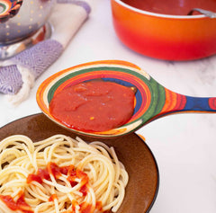 A colorful Baltique® Marrakesh Collection Grand Serving Spoon, 14" from Totally Bamboo holds tomato sauce above a bowl of cooked spaghetti, with some sauce already mixed into the pasta. A red pot and a striped cloth lie in the background on the marble surface.