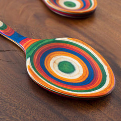 A Baltique® Marrakesh Collection Grand Serving Spoon, 14" from Totally Bamboo, crafted with vibrant multicolored concentric rings in colored birch, sits prominently on the wooden table. Another similar treasure is partially visible in the background.