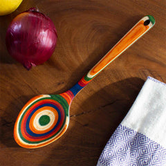 A 14-inch Baltique® Marrakesh Collection Grand Serving Spoon by Totally Bamboo, crafted from colored birch and adorned with striking concentric circles, rests on a wooden table. Next to it are a red onion and a lemon, while a white cloth featuring a blue geometric pattern is partially visible on the right.