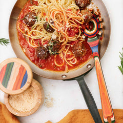 A pan of spaghetti with meatballs in tomato sauce, elegantly topped with grated cheese. Adding a touch of flair, a vibrant Baltique® Marrakesh Collection Spaghetti Spoon by Totally Bamboo rests in the pan. Nearby on a light surface, there's a small wooden bowl filled with cheese and a sprig of rosemary.