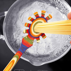 A vibrant spaghetti spoon from the Totally Bamboo Baltique® Marrakesh Collection is being used to portion pasta over a pot of boiling water. This tool, crafted from colorful birch wood in a circular shape with multiple sizing options and adorned with a rainbow pattern, complements the scene beautifully. The pot is positioned on a black stovetop, with bubbles visibly rising in the water.