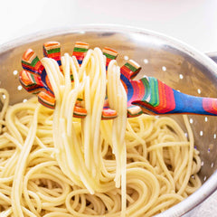 A lively Baltique® Marrakesh Collection Spaghetti Spoon by Totally Bamboo effortlessly lifts a serving of cooked spaghetti over a metal colander filled with more pasta. This colorful, striped birch utensil introduces charm to the bright, well-lit kitchen atmosphere.