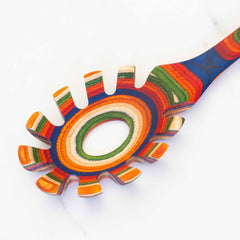 A vibrant spaghetti spoon from the Baltique® Marrakesh Collection by Totally Bamboo, featuring a stunning array of red, blue, green, orange, and yellow stripes crafted from colored birch wood, rests on a white surface. Its distinctive design showcases concentric circles of colors along the handle.