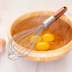 A wooden bowl containing three cracked eggs sits on a marble surface. The 11" Baltique® Marrakesh Collection Balloon Whisk by Totally Bamboo, featuring a colorful wooden handle, rests inside, ready to blend ingredients into a culinary masterpiece.