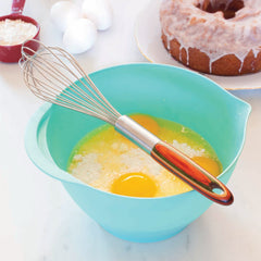 A Totally Bamboo Baltique® Marrakesh Collection Balloon Whisk, with a vibrant wooden handle, rests on a teal mixing bowl filled with eggs, flour, and butter. Nearby, three eggs are placed next to a red measuring cup of flour and a glazed bundt cake on a plate. The entire arrangement sits on an immaculate white countertop.