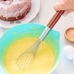 A person is whisking yellow batter using the 11" Baltique® Marrakesh Collection Balloon Whisk by Totally Bamboo, which boasts a colorful wooden handle, all within a light blue bowl. Nearby on the marble surface, there's a glazed bundt cake on a plate, alongside a small bowl of cinnamon and two eggs.