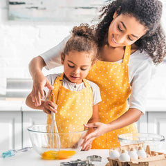 A mother and daughter, wearing matching yellow polka dot aprons, whisk egg yolks with the 11" Baltique® Marrakesh Collection Balloon Whisk by Totally Bamboo, which boasts a colorful wooden handle. They smile as they enjoy their time in the kitchen, surrounded by baking ingredients and cookie cutters.