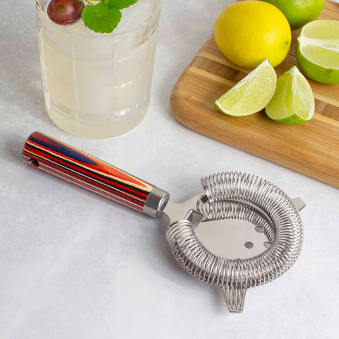 The Baltique® Marrakesh Collection Hawthorne Cocktail Strainer by Totally Bamboo, an essential tool for mixologists, sits on a light surface next to a glass of lemonade garnished with mint and a grape. A wooden cutting board displays whole and sliced citrus fruits, including lemons and limes.