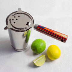 The Baltique® Marrakesh Collection Hawthorne Cocktail Strainer, an essential tool for mixologists from the Totally Bamboo brand, features a vibrant birch handle and rests on a gleaming metal shaker. Beside it are a whole lime, a lemon, and a slice of lime on a light surface.