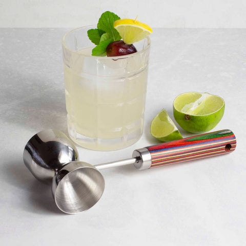 A glass of cocktail with ice, garnished with a lime wedge, mint, and a cherry, sits on the table. Nearby lies an eye-catching muddler from the Marrakesh Collection by Totally Bamboo and a Baltique® Marrakesh Collection Double Jigger made of stainless steel, accompanied by sliced lime wedges.