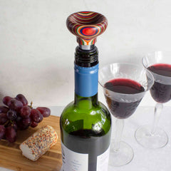 A vibrant Baltique® Marrakesh Collection Bottle Stopper by Totally Bamboo adorns a wine bottle beside two filled glasses. In the background, a cluster of grapes and herb-topped cheese evoke the lively ambiance of Moroccan marketplaces.