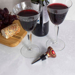 Two glasses of red wine are elegantly displayed on a white surface next to a cheese log on a wooden board. A bottle of wine and the stunning Baltique® Marrakesh Collection Bottle Stopper by Totally Bamboo complete the scene. A bunch of grapes enhances this vibrant arrangement, evoking the colorful marketplaces of Morocco.