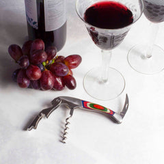 A bunch of red grapes and an open bottle of red wine sit next to two filled glasses on a white surface, accented by the colorful compact design of the Baltique® Marrakesh Collection Waiter's Friend Corkscrew from Totally Bamboo.