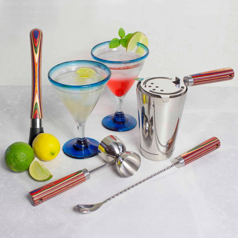 A Baltique® Marrakesh Collection 4-Piece Bartending Kit from Totally Bamboo sits on a marble surface, showcasing vital bartending tools including a muddler, jigger, shaker, and twisted spoon. Blue-rimmed glasses filled with cocktails adorned with lime and mint are encircled by whole and sliced lemons and limes.