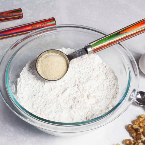 A clear glass bowl filled with white flour is accompanied by a colorful measuring spoon from the Totally Bamboo Baltique® Marrakesh Collection 4-Piece Measuring Spoon Set, poised with sugar above it. Nearby, walnuts and other stylish kitchen decor items rest on the marble countertop.