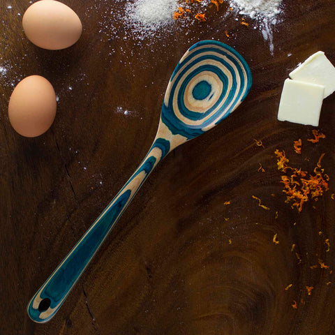 A beautifully patterned cooking spoon from the Totally Bamboo Baltique® Mykonos Collection, featuring blue and natural wood tones, rests on a wooden surface. Nearby are two eggs, butter, flour, and orange zest, suggesting preparation for baking.