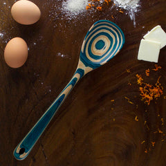 A beautifully patterned cooking spoon from the Totally Bamboo Baltique® Mykonos Collection, featuring blue and natural wood tones, rests on a wooden surface. Nearby are two eggs, butter, flour, and orange zest, suggesting preparation for baking.