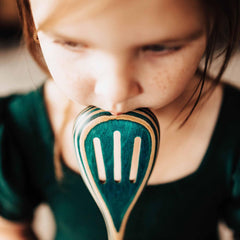 A child with brown hair softly holds a 12-1/2" slotted wooden spoon from the Baltique® Mykonos Collection by Totally Bamboo, featuring an eye-catching design of blue and green stripes, while sporting a dark green shirt. The emphasis is on the spoon's vibrant birch colors held in their mouth.