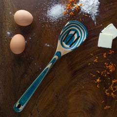 A slotted spoon from the Baltique® Mykonos Collection by Totally Bamboo, featuring a blue swirled design in colored birch, lies on a dark wooden surface. Nearby are two eggs, a small heap of flour, granulated sugar, orange zest, and two pieces of butter.