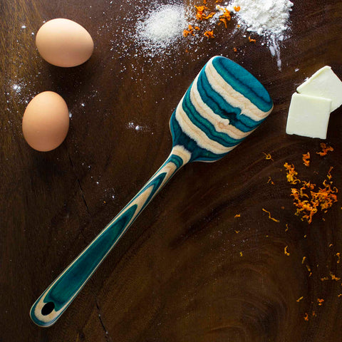 A Totally Bamboo Baltique® Mykonos Collection Spatula, 12-1/2", adorned with blue and beige stripes, rests on a dark wooden surface. Nearby are two eggs, a sprinkle of flour, slices of butter, sugar, and orange zest, suggesting preparations for baking with stylish non-stick cookware.