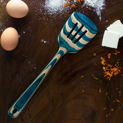 A Totally Bamboo Baltique® Mykonos Collection Slotted Spatula, 12-1/2", in blue and beige rests on a dark wooden surface, surrounded by oranges, flour, butter cubes, and two eggs.